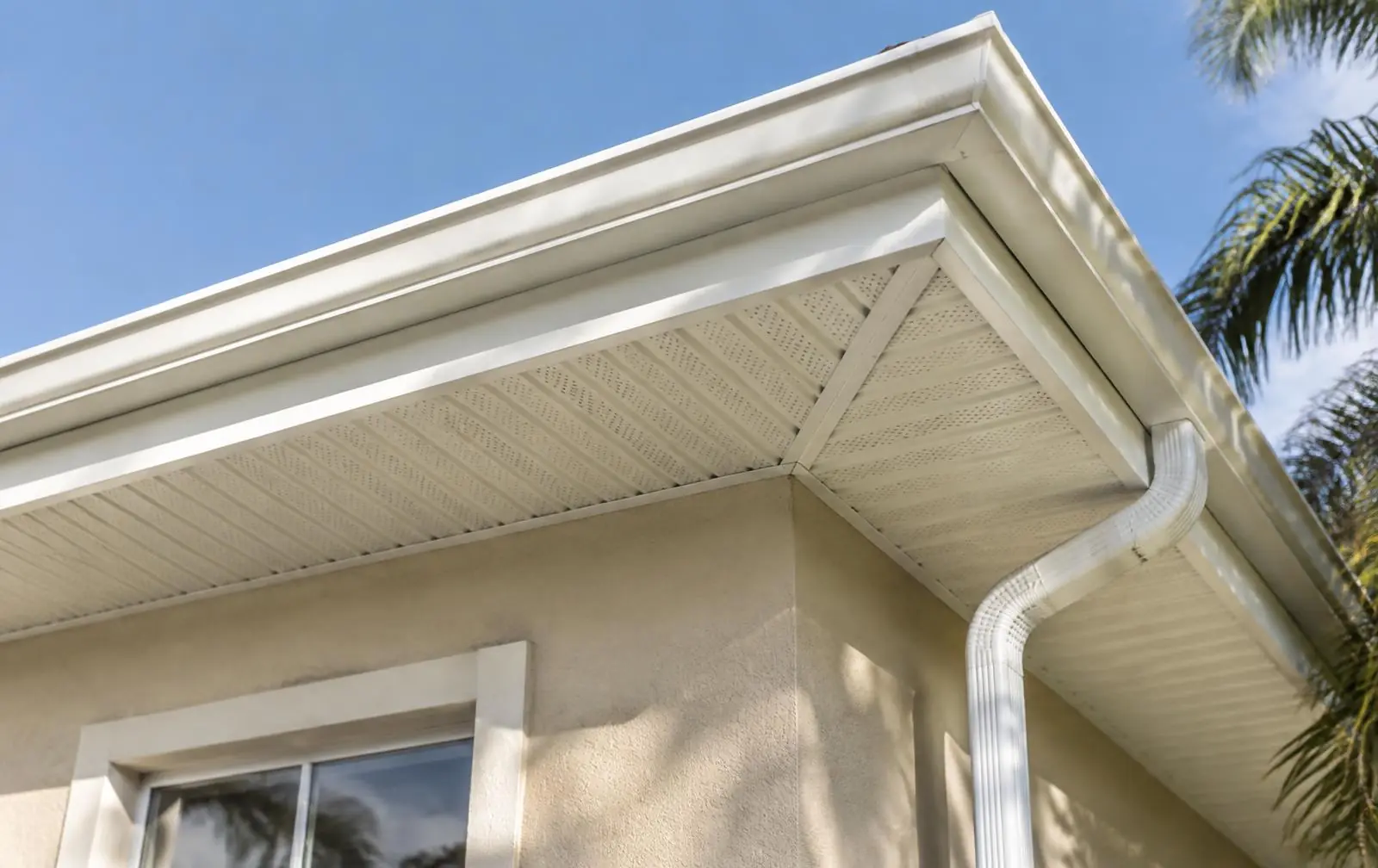 Close-up of white vented soffit panels and fascia with a seamless gutter and downspout on a stucco Florida-style home